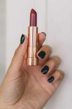 pexels-photo-3060257-3060257 Close-up of a hand with dark polished nails holding a stylish lipstick tube against a neutral background.